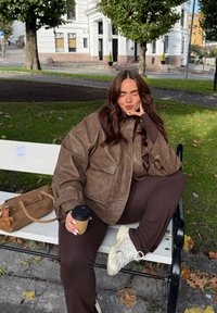 Brązowa, oversizeowa kurtka z eko skóry z dużymi kieszeniami, w zestawie z brązowymi spodniami i białymi trampkami. Trzyma kubek z kawą; obok znajduje się brązowa torebka.