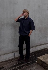 Camicia in denim scuro a maniche corte, vestibilità rilassata, colletto con bottoni, abbinata a pantaloni neri e scarpe marroni. Sfondo di una parete in cemento chiaro.