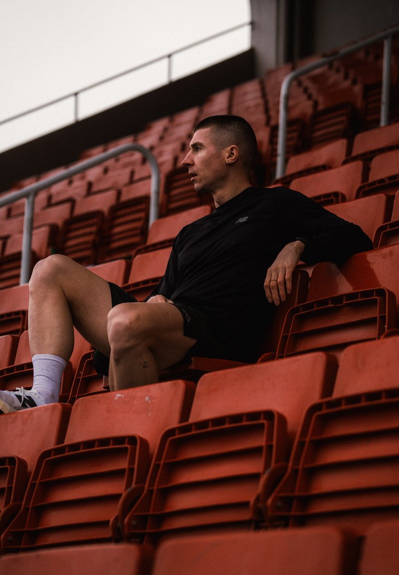 Άνδρας με μαύρα αθλητικά ρούχα που κάθεται μόνος σε κόκκινα καθίσματα γηπέδου, κοιτάζοντας προς τα πλάγια με το ένα χέρι να ακουμπάει στην πλάτη του καθίσματος.
