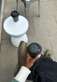 Tazze di caffè su un tavolo rotondo bianco, una mano che tiene una tazza nera, scarpe marroni chiare con cuciture gialle e pavimentazione grigia.