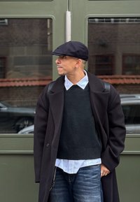 Fekete felöltő sötét pulóver fölött, fehér inggallér kilátszik, kék farmernadrág, és fekete lapos sapka, zöld ajtók előtt állva.