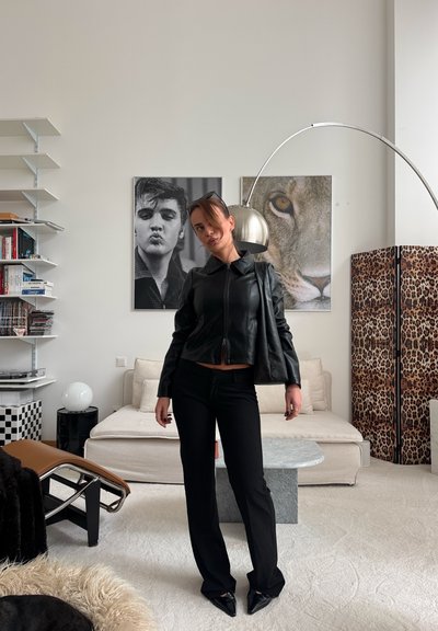 Mujer con chaqueta y pantalones de cuero negros posa en una sala de estar moderna con retratos de león y hombre, separador de ambientes con estampado de leopardo y lámpara de pie arqueada.