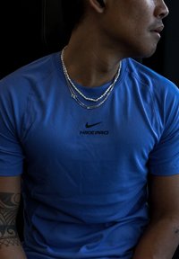 Nike Pro blå atletisk skjorte med korte ærmer, en tætsiddende pasform og en sort logo i midten. Flere lagdelte halskæder tilføjer detaljer.