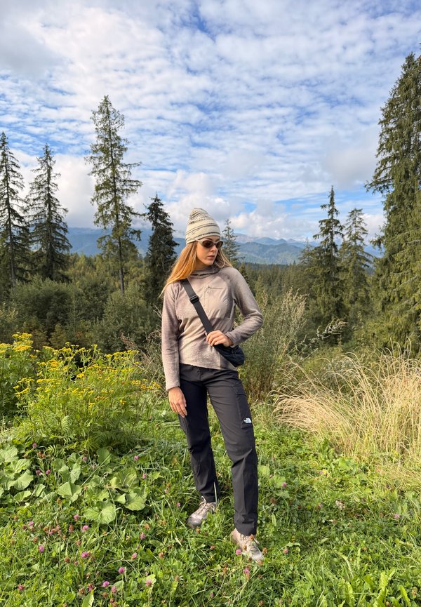 Una mujer vestida para hacer senderismo está entre flores silvestres en un sendero de montaña, con árboles de hoja perenne y montañas lejanas a sus espaldas.