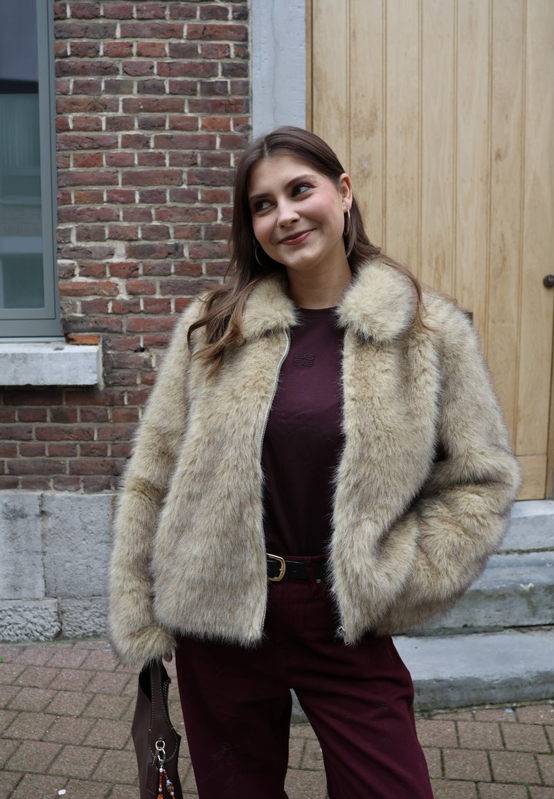 Veste en fausse fourrure beige à la texture douce, fermeture éclair à l'avant et col, associée à un haut bordeaux et un pantalon assorti. Sac à main marron visible.