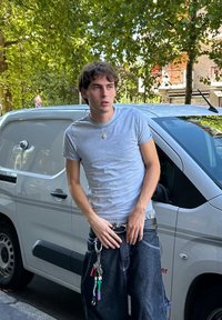 T-shirt cinza, calças baggy de denim escuro, e um cinto preto com um porta-chaves anexado. O fundo apresenta uma furgão prateada e árvores verdes.