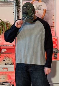 Personne portant un pull côtelé bicolore et une casquette camouflage, ajustant la casquette en se tenant dans une pièce avec des baskets et des étagères décoratives en arrière-plan.