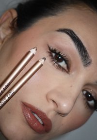 ZweiMake-up-Bleistifte mit metallic-goldener Verpackung, einer in heller und einer in dunklerem Farbton, werden in der Nähe eines Gesichts mit neutralem Make-up gehalten.