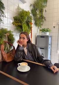 Femme aux cheveux longs portant une veste en cuir et une chemise à carreaux assise à une table avec un café, entourée de couronnes vertes suspendues et de plantes.