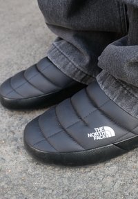 Juodai pūkuoti šlepetės su siuvinėtu raštu ir guminiais padais. Šone yra balta "The North Face" logotipo emblema.