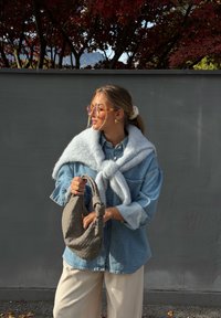 Denimist särk, kaelal seotud halli kudumitriibuga sall, kreemjad laia lõikega püksid, käes tekstureeritud hall käekott. Taustal on punased lehed.