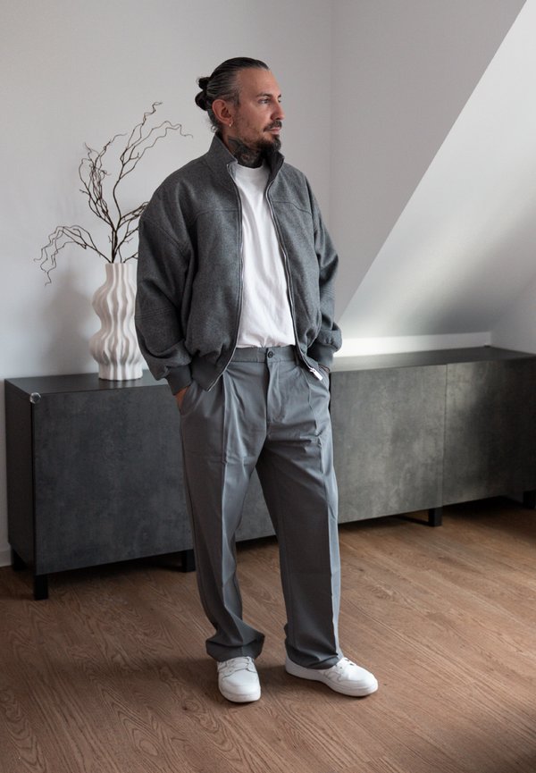 Un homme barbu aux cheveux attachés en arrière se tient debout, les mains dans les poches. Il porte une veste et un pantalon gris ainsi que des baskets blanches, dans une pièce moderne.