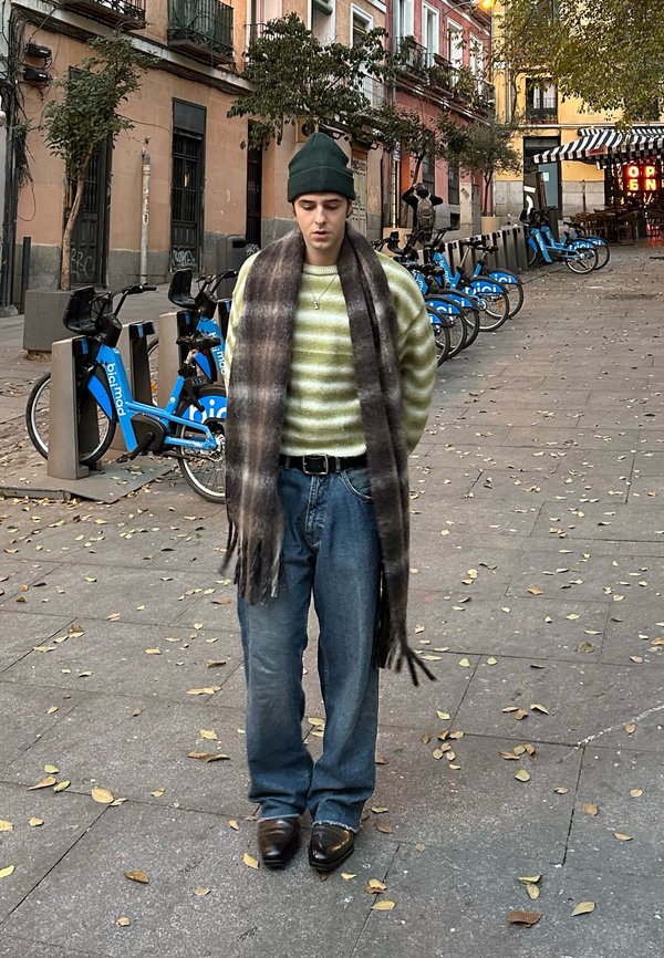 Grøn og creme stribet sweater, grå plaid tørklæde, blå jeans og blanke sko. Udendørs miljø med cykler og faldne blade i baggrunden.