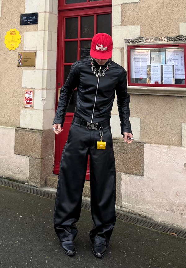 Črn usnjen zip-up zgornji del in široke hlače. Dodatki vključujejo rdečo kapo in rumeno torbico na ključ. Okolica vključuje teksturirane kamnite stene.