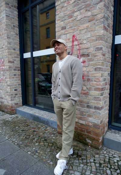 Cardigan de canalé beige, camiseta blanca y pantalones caqui claros. Zapatillas blancas. De pie junto a una pared de ladrillo con ventanas.