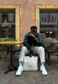 Weisse, gesteppte Tragetasche, schwarze Lederjacke, runde gelbe Tisch und weisse Sneakers. Der Hintergrund zeigt Schaufenster und dekorative Lichter.