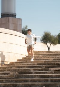 Witte sport t-shirt en beige korte broek gedragen door een persoon die van stenen trappen naar beneden rent. De achtergrond toont groen en een stenen muur.