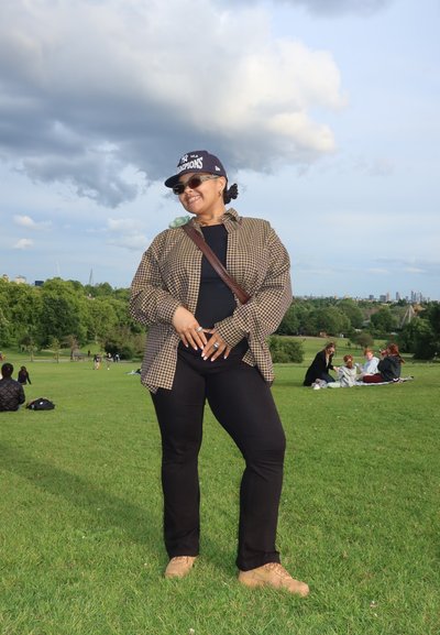 Persona vestida con una camiseta negra de cuello redondo, una camisa de cuadro de gran tamaño, pantalones negros y zapatos beige, posando sobre hierba con un fondo de parque.