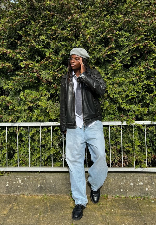 Zwarte leren jas, grijze baret, witte shirt, grijze das, lichtblauwe baggy jeans, zwarte schoenen; staand bij groenvoorziening en een metalen leuning.