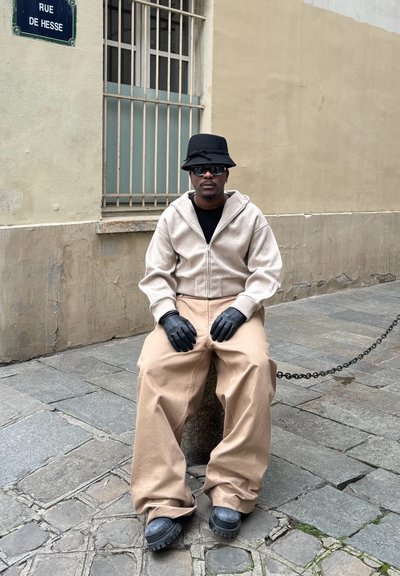 Sudadera beige con cremallera, camiseta negra, pantalones beige sueltos, guantes negros y un sombrero de cubo negro. Sentado en una piedra con una cadena de fondo.