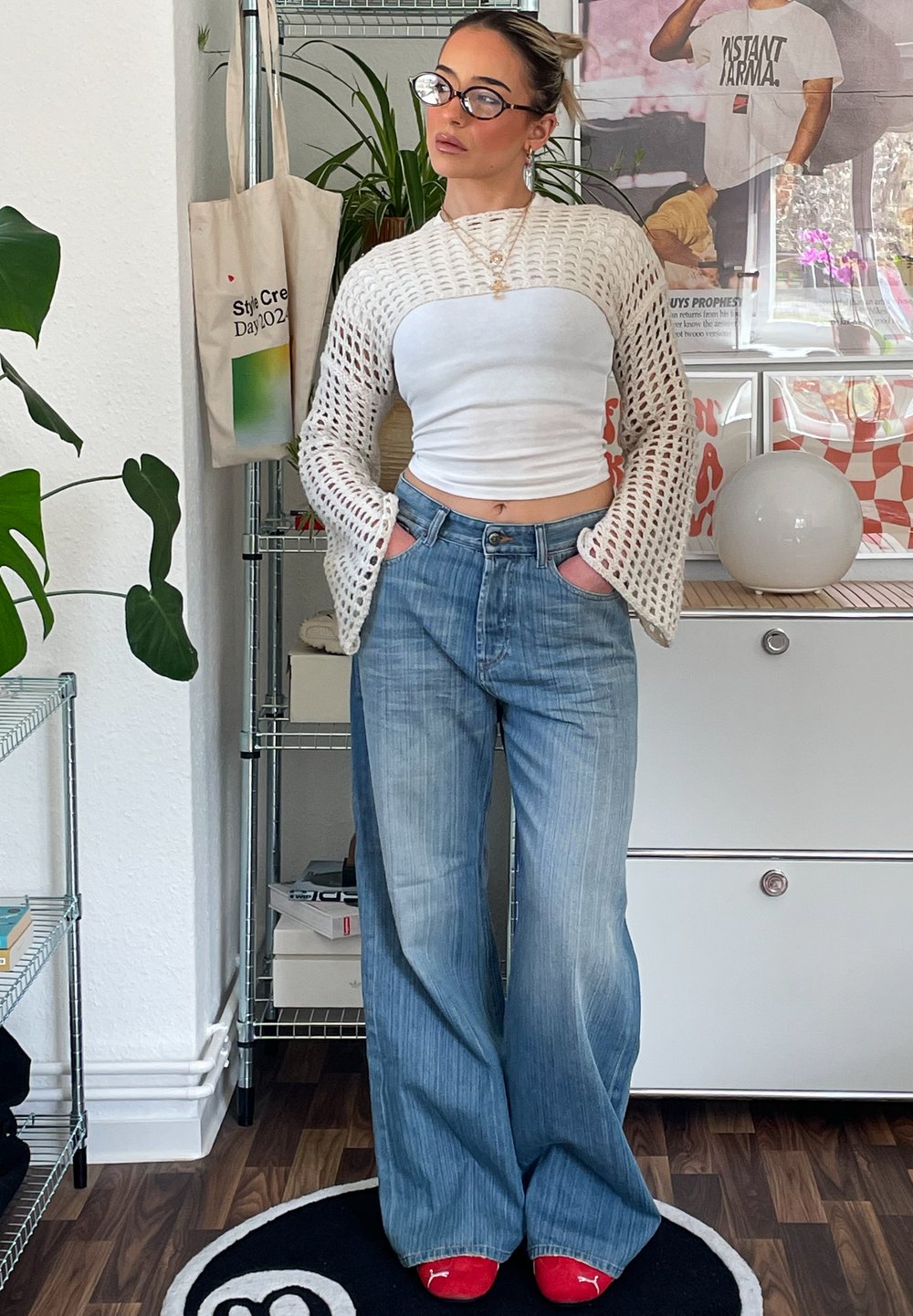 Top curto de malha branca com mangas largas, jeans de cintura alta azul claro e sapatos vermelhos sem fecho. Ambiente interior com plantas e itens decorativos.