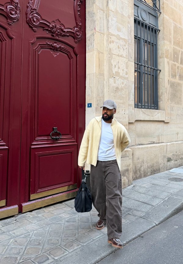 Egy férfi kockás sapkában, krém színű dzsekiben, fehér ingben és barna nadrágban, aki kőburkolatos járdán sétál el egy magas vörös ajtó és egy bézs kőépület mellett.