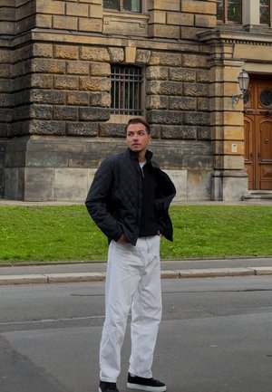 Chaqueta negra acolchada sobre un suéter negro, pantalones holgados blancos y zapatillas negras, con un edificio de piedra y un césped verde de fondo.