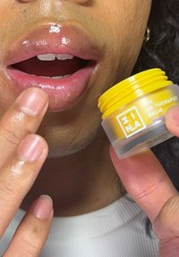 Keltainen huulivoide kirkkaassa purkissa, jota pidetään lähellä kiiltäviä huulia. Henkilön sormet levittävät voiteen huulilleen, näyttäen sileän koostumuksen.