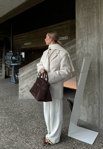 Abrigo texturizado beige, cuello alto blanco, pantalones crema y bolso marrón con asas. Zapatillas oscuras con cordones amarillos. Fondo de concreto.