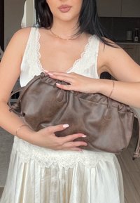 Vrouw in een wit jurk met kant afgewerkt, die een grote bruine leren tas tegen haar borst houdt, met verfijnde gouden sieraden om haar polsen en nek.