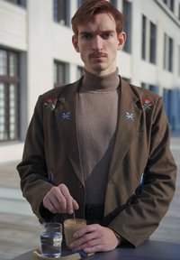 Blazer marrón con bordado floral y de lazo, suéter de cuello alto gris, sosteniendo un vaso de café helado y un vaso de agua, fondo minimalista.