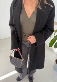 Cappotto di lana nero sopra una maglia testurizzata verde oliva, pantaloni grigi a gamba larga e una borsa con motivo e tracolla marrone. Calzature nere lisce.