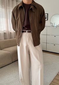 Veste légère marron avec fermeture éclair et boutons-pression, associée à un pantalon beige à jambes larges. Le tissu texturé ajoute un intérêt visuel.