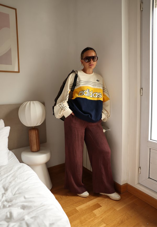 Mujer con gafas de sol grandes, una sudadera Adidas amarilla, azul y blanca, pantalones anchos marrones y zapatos blancos, de pie dentro de una habitación junto a una ventana y una cama.
