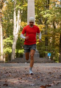 Juoksija punaisessa lyhythihaisessa paidassa ja harmaissa shortseissa, päässään valkoinen pipo ja jalassaan vaaleansiniset lenkkarit, lehtien peittämällä polulla puistossa.