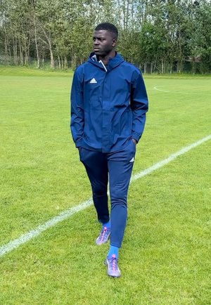 Hombre con chándal azul de Adidas caminando sobre el césped cerca de la línea límite del campo de fútbol, con árboles al fondo, con las manos en los bolsillos, mirando hacia la izquierda.