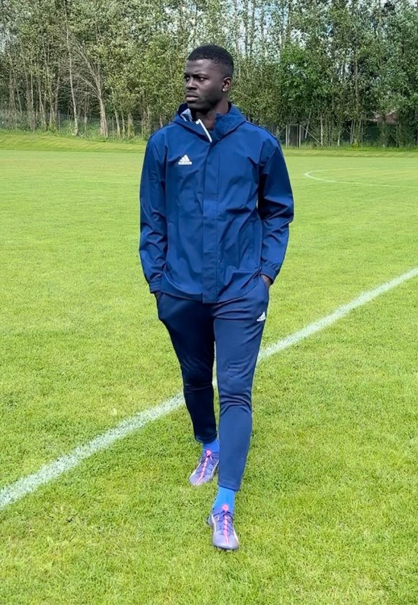 Muž v modrej Adidas teplákovej súprave kráča po tráve pri hranici futbalového ihriska s množstvom stromov v pozadí, ruky v vreckách, pozerá sa doľava.