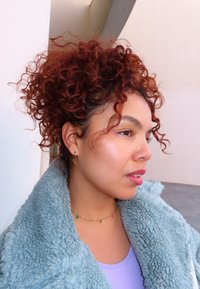 Femme aux cheveux roux bouclés portant un manteau bleu clair duveteux et un collier de perles colorées, tournée vers la droite devant un mur pâle.