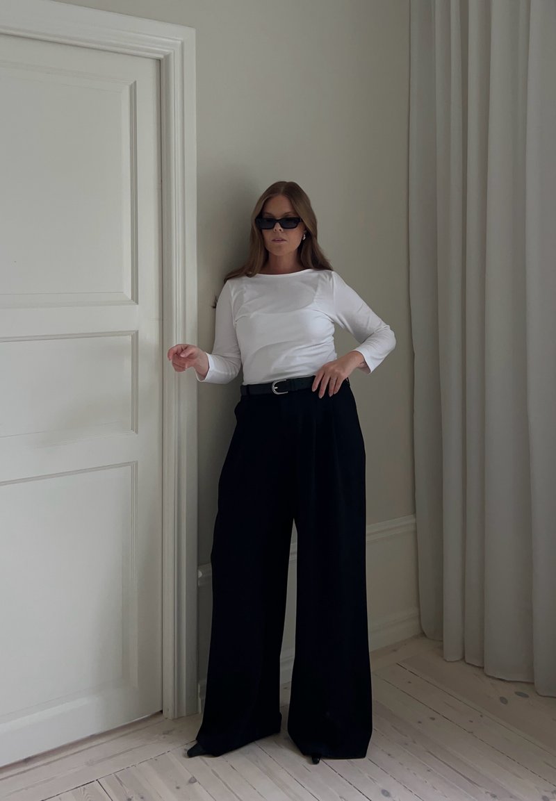 Femme portant un pantalon noir à jambes larges, un haut blanc à manches longues, une ceinture noire et des lunettes de soleil foncées, debout contre un mur clair près d'une porte.