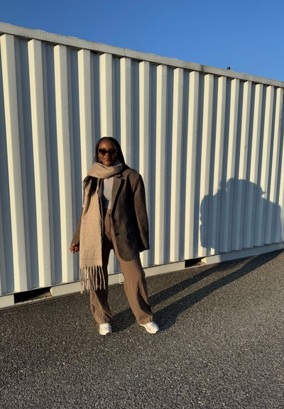 Blazer marrón, suéter de cuello alto gris, bufanda marrón claro, pantalones marrón claro y zapatillas blancas. De pie contra una pared de rayas verticales, en un entorno iluminado por el sol.