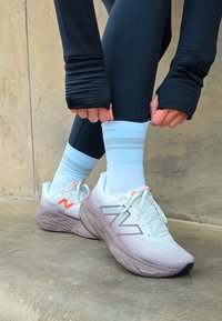 Käed reguleerivad valgeid sokke valgete ja roosa New Balance'i jooksujalatsite kohal betoonpinnal beeži seina lähedal.