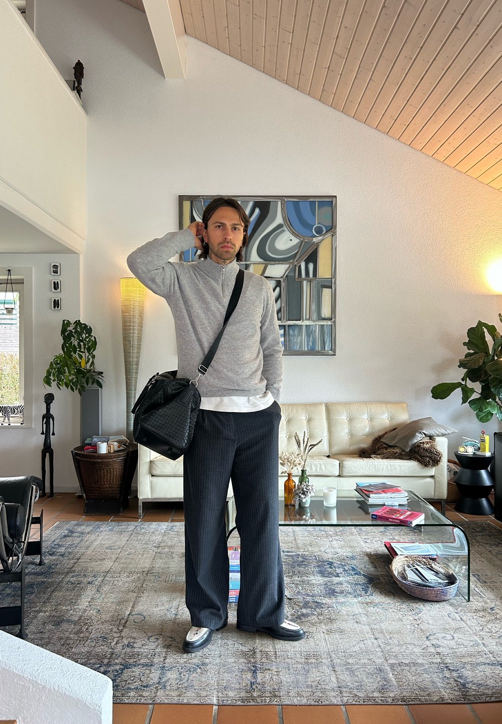 Tenue décontractée comprenant un pull zippé gris, un sac noir, un pantalon à rayures taille large et des chaussures noires. En arrière-plan, un canapé crème et des œuvres d'art.