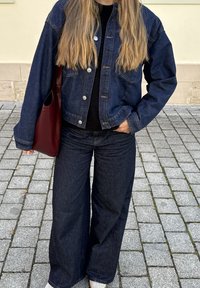 Traper jakna tamno plave boje, u kombinaciji s crnim puloverom i širokim tamnim farmerkama. Burgađeva torba nošena je preko ramena.