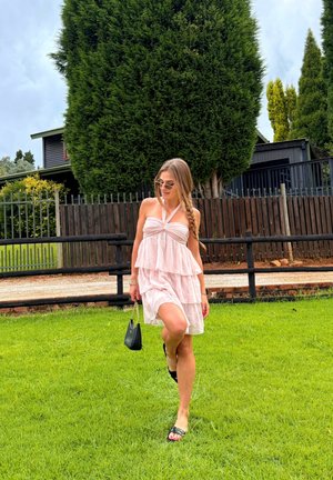 Mujer joven con vestido rosa de capas y gafas de sol caminando sobre césped verde cerca de una cerca de madera y árboles altos, sosteniendo un bolso pequeño negro.