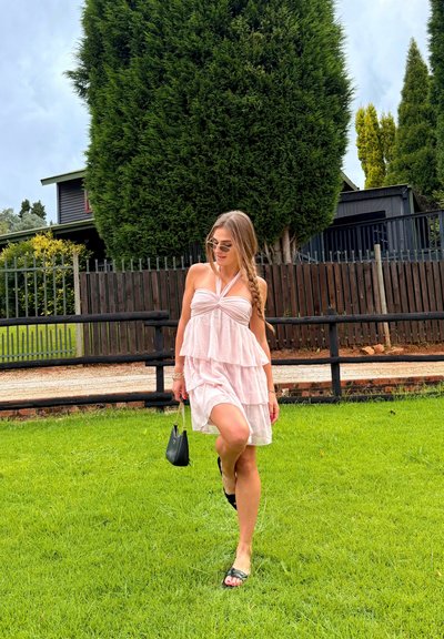Mujer joven con vestido rosa de capas y gafas de sol caminando sobre césped verde cerca de una cerca de madera y árboles altos, sosteniendo un bolso pequeño negro.