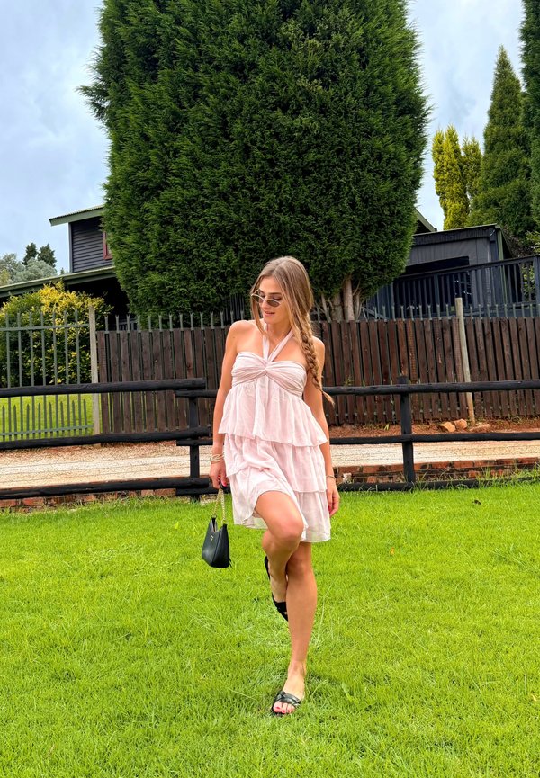 Noor naine roosas kihilises kleidis ja päikeseprillidega kõndimas rohelisel muruplatsil puidust aia ning kõrgete puude lähedal, käes väike must käekott.