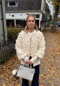 Krémový kardigan s texturou a chlupatým vzorem, nošený přes béžový top, doplněný šedou kabelkou. V pozadí jsou podzimní listy.