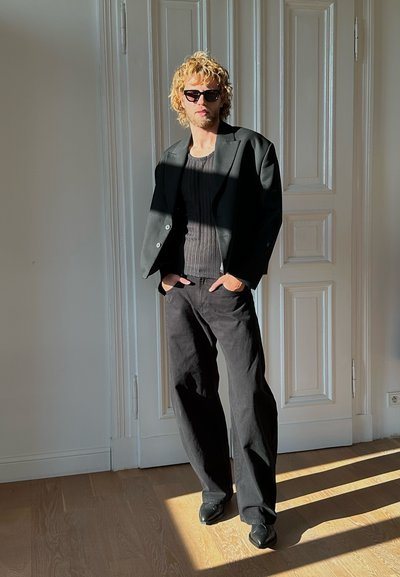 Blazer oversize negro, top negro acanalado y pantalones negros de pierna ancha. Estilizado con gafas de sol negras y zapatos elegantes. Una luz brillante proyecta sombra.