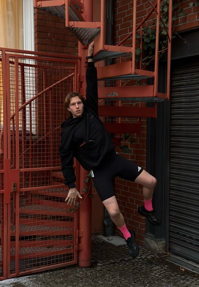 Joven con ropa deportiva negra y calcetines rosa brillante colgado de una escalera de caracol metálica roja en el exterior de un edificio de ladrillo.