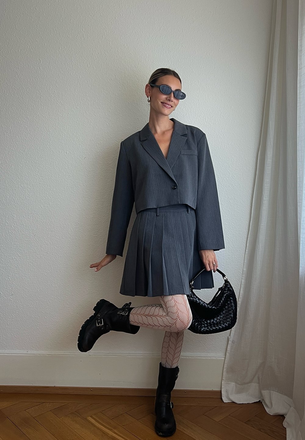 Conjunto cinza ajustado de duas peças com um blazer curto e uma saia plissada. Meias-calças de rede transparentes, botas pretas de tornozelo e uma bolsa preta em tecido trançado.